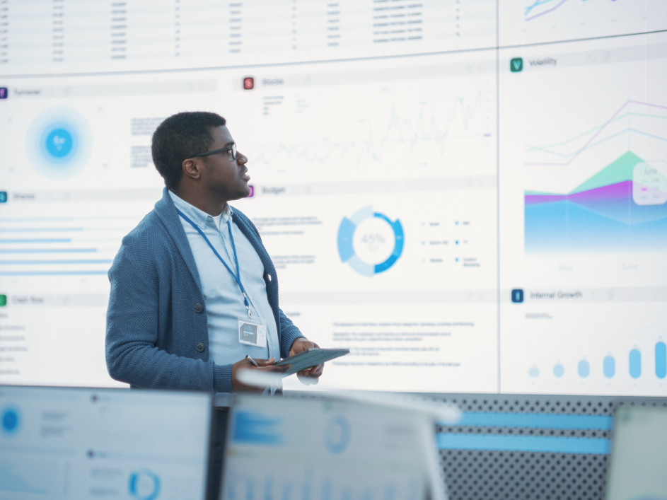 Black male general manager presenting data on a large digital screen to a diverse group of consulting company employees in a monitoring room. Colleagues are seated at desktop computers, attentively listening.