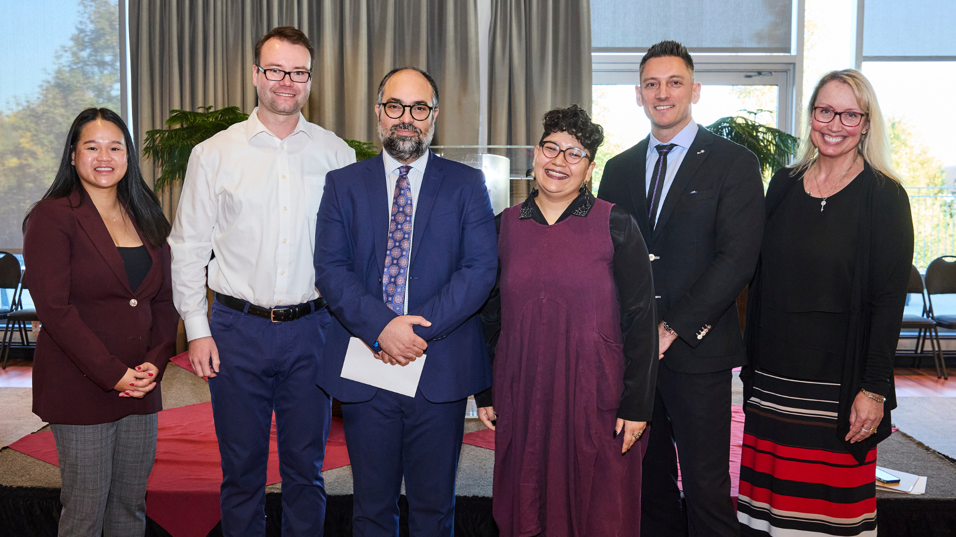 Six DeGroote faculty members pose together, smiling in front of a podium.