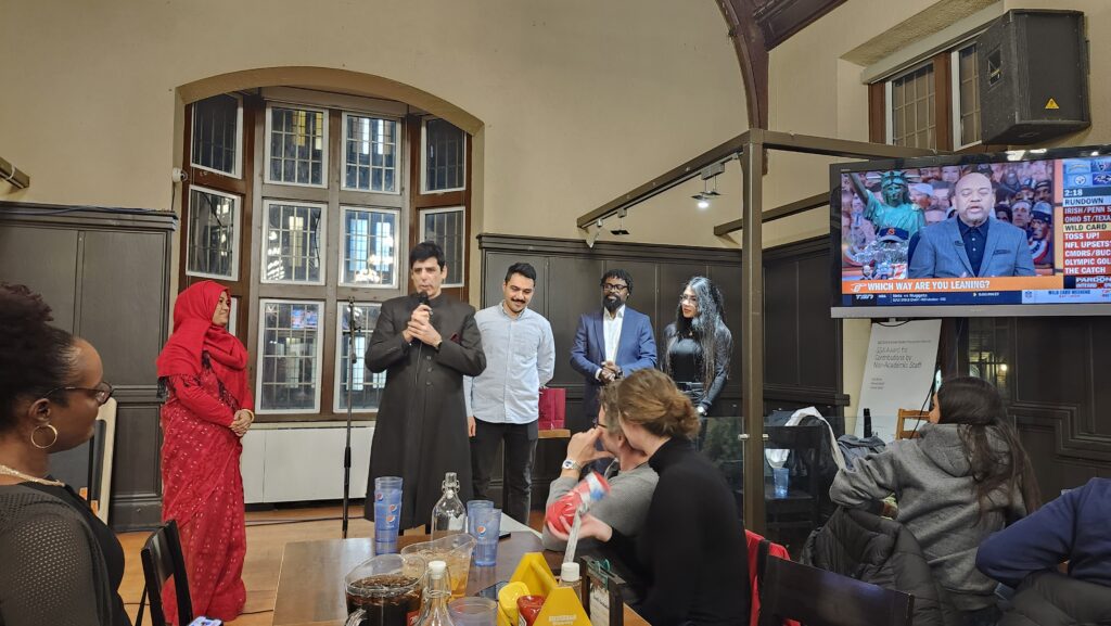 A man speaks to his colleagues in a room with a television during a dinner gathering.