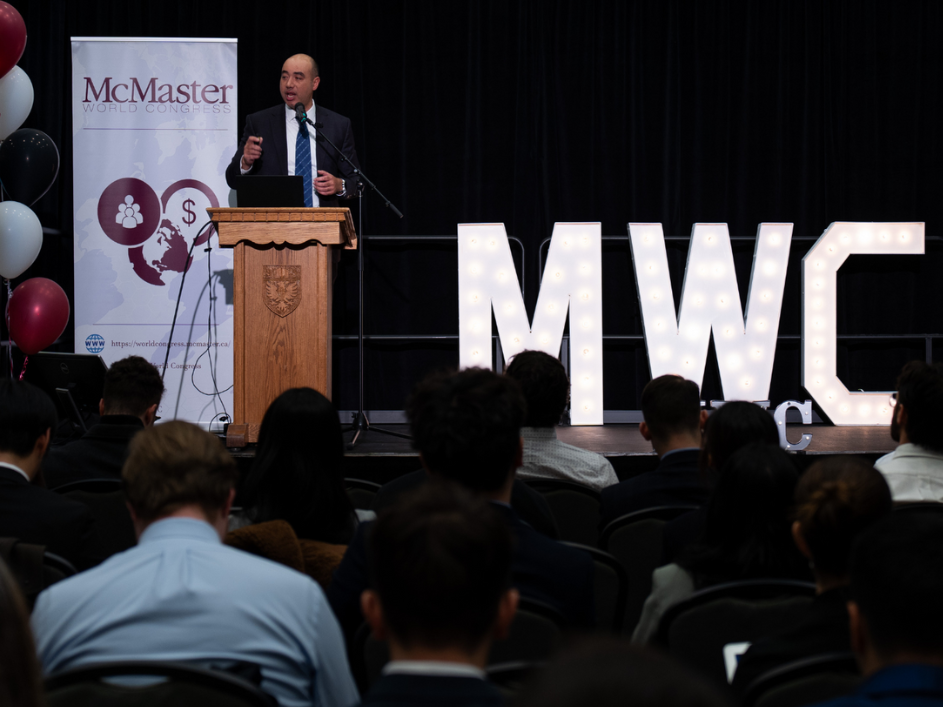 A speaker addressing an audience from the podium at the McMaster World Congress. The stage is adorned with the MWC logo.