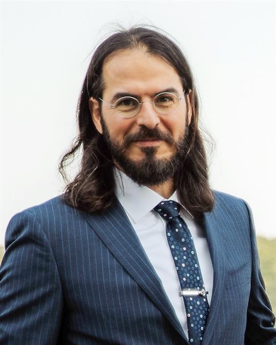 Headshot of Keiwan Wind, smiling man with long, dark hair, glasses, wearing a navy suit.