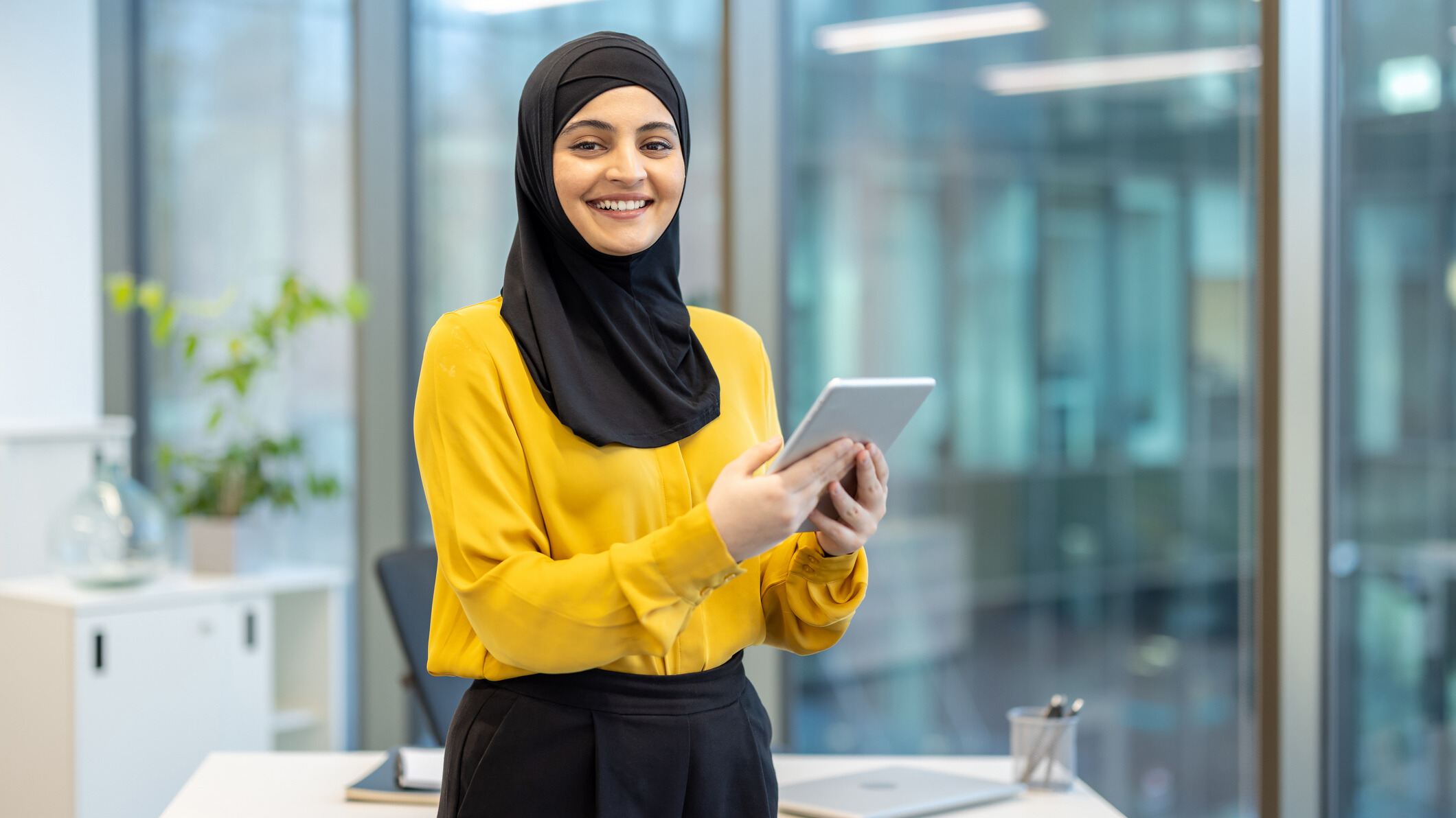 a woman in black hijab and bright yellow blouse stands smiling in an office while holding an ipad