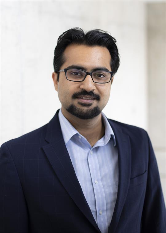 Headshot of Zohair Alam, a smiling man with a beard, dark hair, glasses, wearing a black suit.