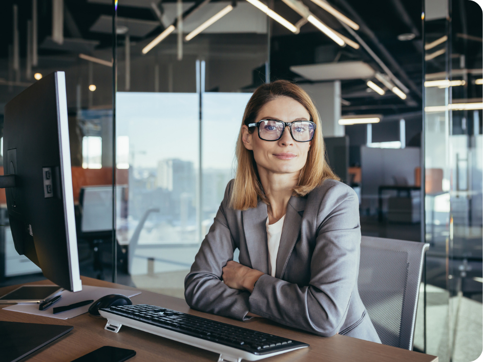 A blond woman with glasses and wearing a gray blazer smiling while sitting on a desk in front of a computer.