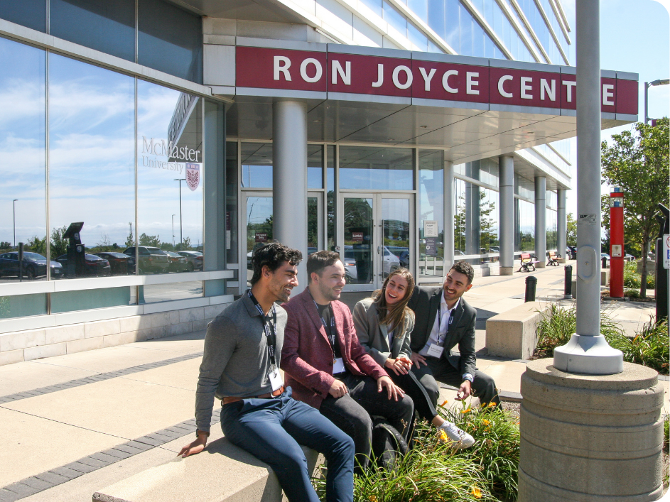 Four MBA students sitting outside of the Ron Joyce Centre having a conversation and smiling.