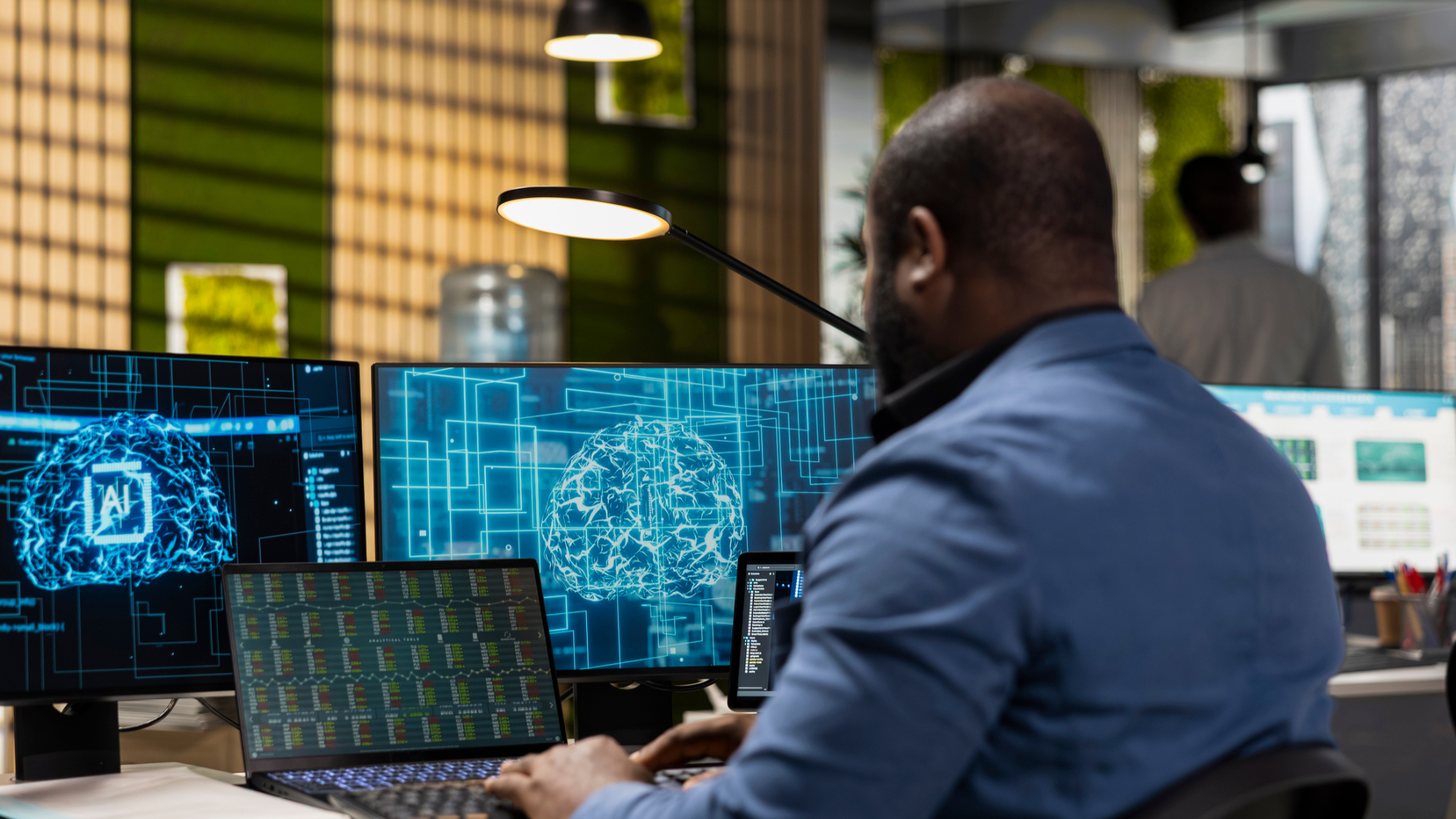 a man turned to a desk with multiple screens, working on a laptop, wearing a blue button up.